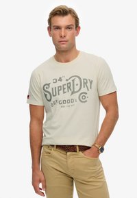  camiseta de color crema con una gran impresión gráfica gris que dice "SuperDry". Combinada con pantalones caqui y un cinturón de cuero trenzado marrón.