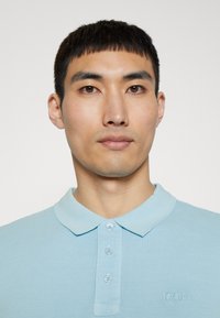 Jasnoniebieska koszulka polo wykonana z teksturowanej tkaniny, z kołnierzykiem i trzema guzikami. Logo haftowane na klatce piersiowej.