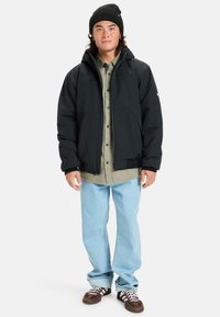 Jeune homme portant un bonnet noir, une veste noire sur une chemise grise, un jean bleu clair et des baskets brunes, se tenant devant un fond blanc.