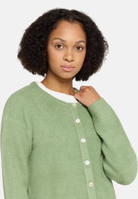Zielony dzianinowy kardigan z okrągłym dekoltem, zapinany na guziki z przodu, z prążkowanymi mankietami. Teksturowana tkanina dodaje miękkości i ciepła.