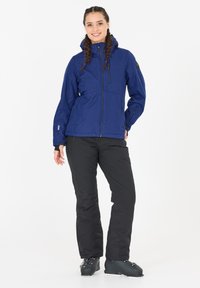 Marineblaue wasserdichte Jacke mit Kapuze, durchgehendem Reißverschluss und verstellbaren Bündchen. Kombiniert mit schwarzen Hosen und schwarzen Skistiefeln.