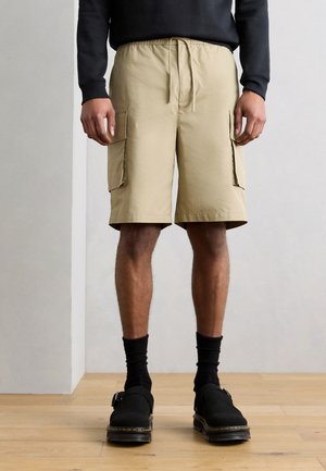 Άνδρας που φοράει μπεζ cargo σορτς με πλαϊνές τσέπες, μαύρες κάλτσες, μαύρα slip-on παπούτσια και μακρυμάνικη μαύρη μπλούζα, στέκεται σε ξύλινο πάτωμα.