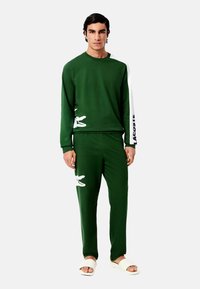 Sötétzöld pulóver és nadrág szett puha anyagból. Fehér Lacoste logóval és krokodil grafikával; lazán illeszkedő, hosszú ujjú, egyszerű dizájn.