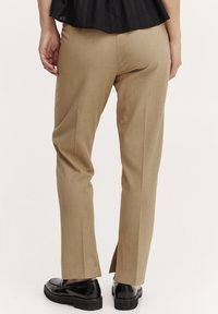 Pantaloni beige su misura con una texture liscia, vestibilità slim e spacco alle caviglie; abbinati a mocassini neri spessi.