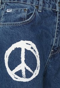Jeans de ganga com lavagem azul escura, apresentando um grande símbolo da paz branco pintado na parte externa da coxa esquerda. Detalhes de costura e ferragens visíveis.