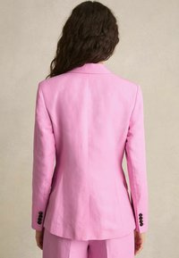 Růžový přizpůsobený blazer s kulatými klopami, strukturovanými rameny a přiléhavým střihem. Obsahuje černé knoflíkové detaily na rukávech.