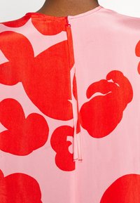 Patrón floral rojo sobre tejido rosa, con un cierre de cremallera en la parte posterior. El diseño incluye formas grandes y suaves con una textura suave.