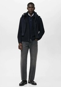 Sudadera con cremallera azul marino sobre una camiseta azul marino, jeans grises y zapatos negros. La sudadera presenta un cuello texturizado y un diseño de ajuste relajado.