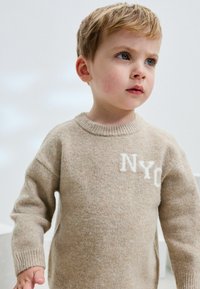 Bež vuneni pulover s okruglim izrezom, s bijelim natpisom "NYC" na prednjoj strani. Tekstura izgleda mekana i pletiva, s rebrastim rukavima.