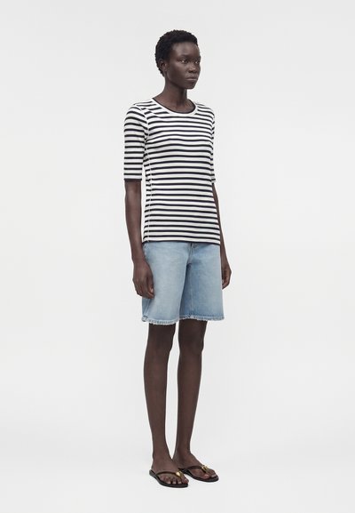 Γυναίκα που φοράει ριγέ μαύρο και λευκό κοντομάνικο πουκάμισο, ανοιχτό μπλε denim σορτς μέχρι το γόνατο και μαύρα σανδάλια με χρυσές λεπτομέρειες.