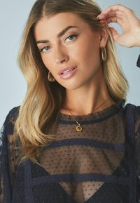 Blouse en tissu transparent bleu marine avec un motif à pois et un bord en dentelle. Collier pendentif carré en or et boucles d'oreilles créoles en or assorties. Tissu doux et texturé.