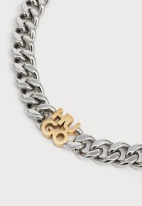 Metallkedja i halsband med växlande blanka silverlänkar och en gyllene "HUGO"-logocharm, med en platt, elegant design och slät textur.