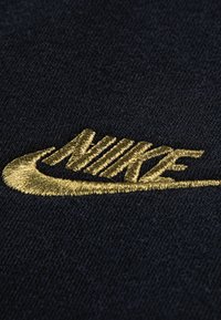 Nike logo zelta izšuvumā uz melna auduma fona. Dizainā ir gludas līknes un asu malu kombinācija, kas radī dinamisku izskatu.