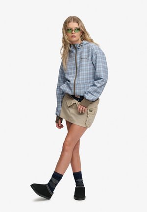 Ung kvinde iført grønne solbriller, en blå ternet lynlåstrøje, beige mini nederdel, mørke mønstrede strømper og sorte slip-on sko.