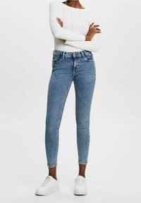 Jeans ajustados de mezclilla azul claro, con cintura alta, bolsillos frontales, acentos desteñidos, combinados con zapatillas blancas y un suéter blanco acanalado.