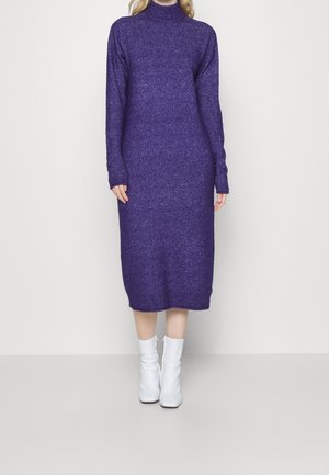 Robe midi en maille violette avec un col montant, des manches longues et une coupe ample. Associée à des bottines blanches. Texture lisse, sans motifs visibles.