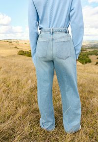 Svetlo modré vysoké nohavice s širokými nohavicami, dvomi zadnými vreckami a klasickou denimovou textúrou, umiestnené na trávnatom pozadí.