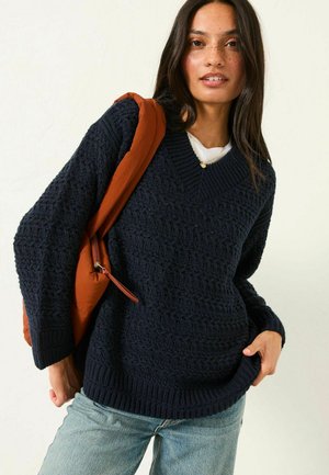 Navy blue knitted sweater with a V-neck, textured pattern, ribbed cuffs and hem, paired with light blue jeans and an orange bag.