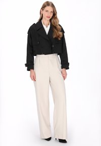 Junge Frau mit langen, welligen Haaren trägt eine schwarze, verkürzte zweireihige Jacke, ein weißes Hemd, beige Hosen mit weitem Bein und schwarze High Heels, stehend.