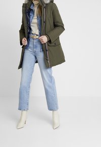 Olivově zelená parka s kapucí lemovanou kožešinou, zapínáním na zip a knoflíky, s bočními kapsami, nošená přes džínovou košili a světlé modré džíny s vysokým pasem.