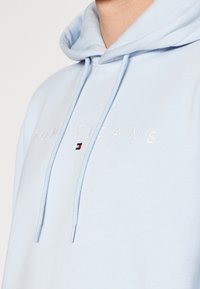 Světlé modrá mikina s kapucí z měkké látky, s šňůrkami a vyšitým logem "TOMMYJEANS" s červenými, bílými a modrými detaily.