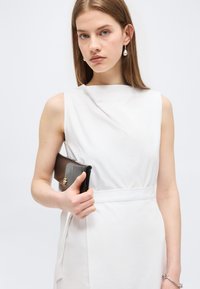 Jeune femme en robe blanche sans manches tenant une pochette noire, portant des boucles d'oreilles pendantes en perles et un bracelet argenté, sur fond uni.