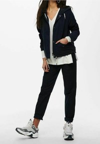 Giacca a vento blu con cappuccio e zip argentate, indossata sopra una maglietta grigia chiara, abbinata a pantaloni scuri e sneakers bianche con dettagli grigi.