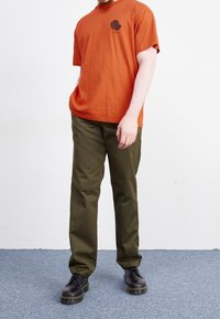 Hombre vestido con una camiseta gráfica naranja, pantalones verdes oliva y botas negras de cordones, de pie sobre una alfombra gris contra una pared blanca.