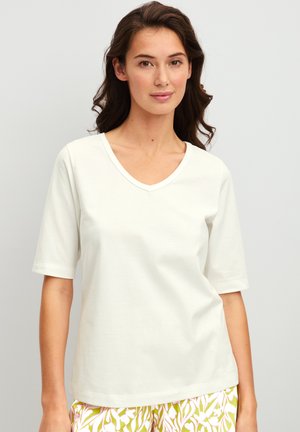 Femme aux longs cheveux foncés portant un t-shirt blanc uni à col en V et manches courtes ainsi qu’un pantalon jaune à motifs sur un fond clair.