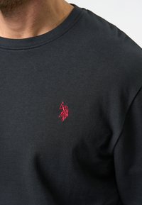 Tricou negru cu guler rotund, din bumbac, având un mic logo brodat roșu în zona stângă a pieptului. Textură netedă, greutate medie.
