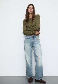 Olivno zeleni pleteni pulover, svetlo modri široki traper hlače z zbledelim spodnjim delom in rjave semiš čevlje. Manekenka stoji z rokami prekriženimi.