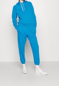 Blå bomullshoodie med en framficka, i kombination med matchande joggingbyxor. Har elastiska muddkanter och midjeband. Vita sneakers som accent.