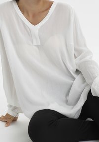 Chemisier blanc à manches longues avec un col en V et une texture subtile, associé à des leggings noirs. Le chemisier a une coupe ample et des manches retroussées.