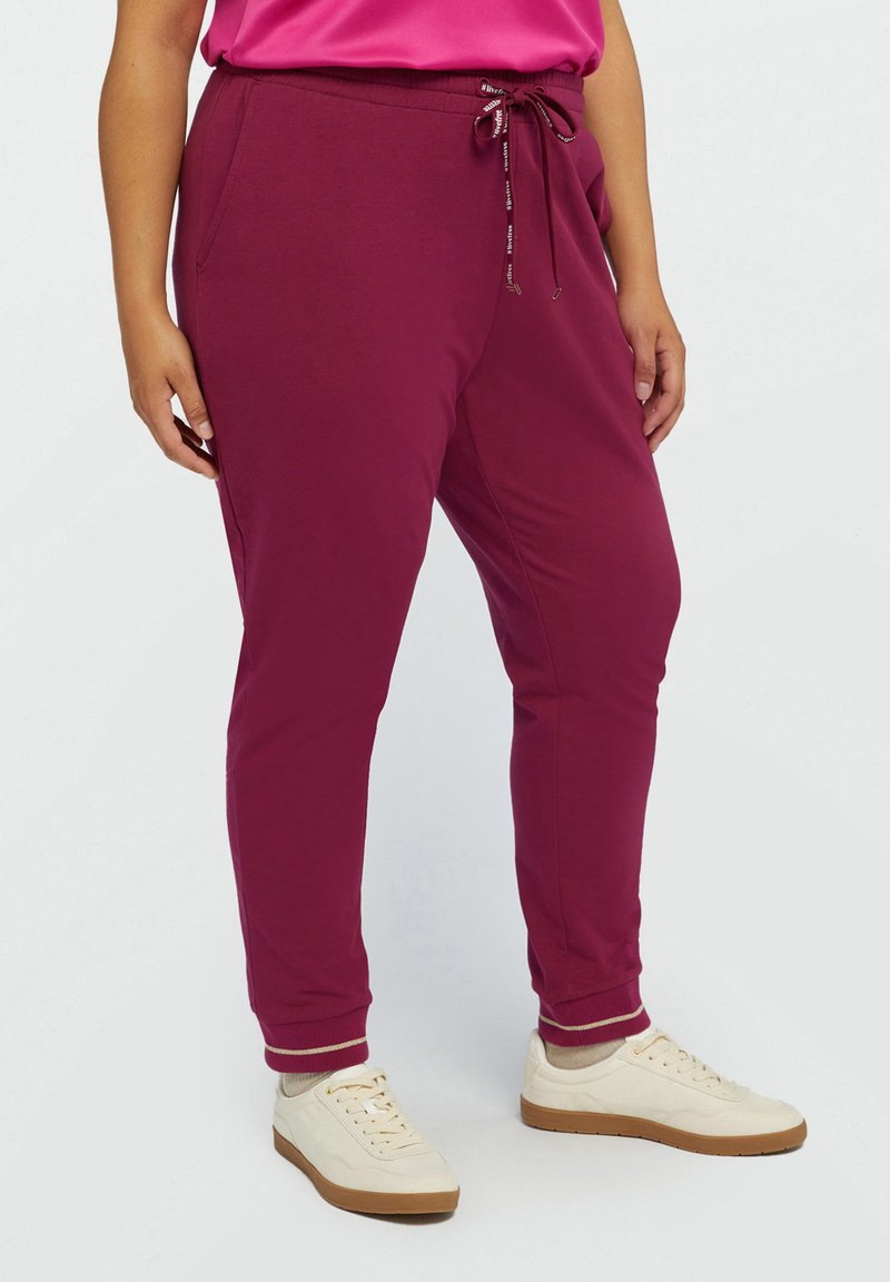 Pantaloni de jogger burgundy cu talie ajustabilă cu șnur, buzunare laterale și manșete ribbate, confecționați dintr-un material moale, asortați cu sneakers crem.