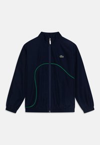 Chaqueta de cremallera azul marino con puños elásticos, diseño de línea curva verde y pequeño logo de cocodrilo verde en el lado izquierdo del pecho.