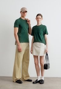 Δύο άτομα φορούν τα ίδια σκούρα πράσινα μπλουζάκια. Το ένα συνδυάζει με μπεζ φαρδιά παντελόνια και καπέλο, το άλλο με λευκή φούστα και μαύρη τσάντα.