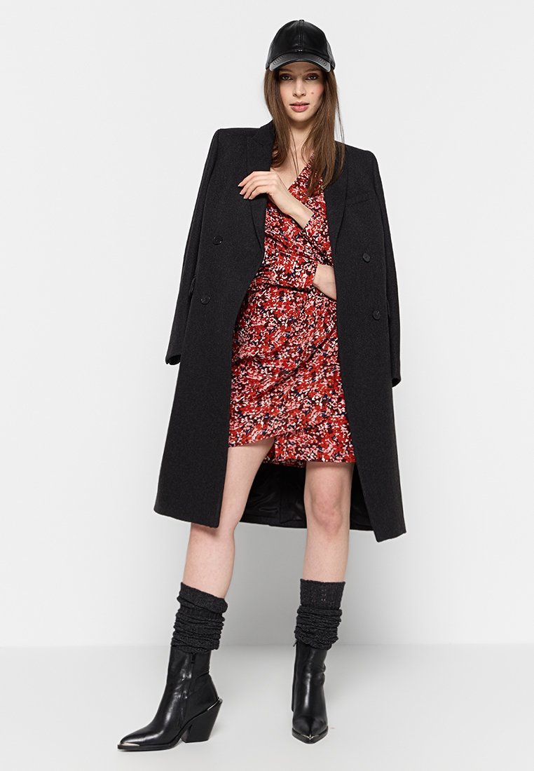 Manteau noir à double boutonnage sur une robe fleurie rouge, associé à une casquette en cuir noir et des chaussettes noires lâches rentrées dans des bottes à talons.