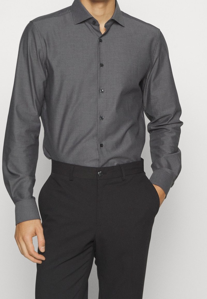 Chemise gris foncé à manches longues avec un petit motif à points, dotée d'un col boutonné et de boutons noirs ; associée à un pantalon noir.