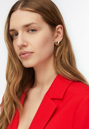 Giovane donna con lunghi capelli castano chiaro, che indossa orecchini dorati a forma di cuore e un blazer rosso acceso, che guarda leggermente di lato.