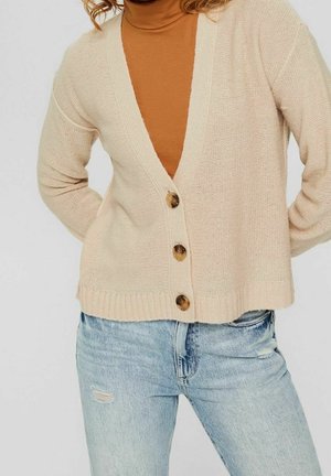 Cárdigan de punto beige con un escote en V profundo, que presenta cuatro botones de madera y un dobladillo acanalado, superpuesto a un cuello de tortuga marrón y jeans azules.