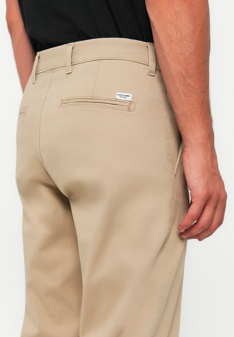 Pantalons en coton beige avec une coupe slim, comportant des poches arrière, des coutures discrètes et une petite étiquette sur la ceinture.