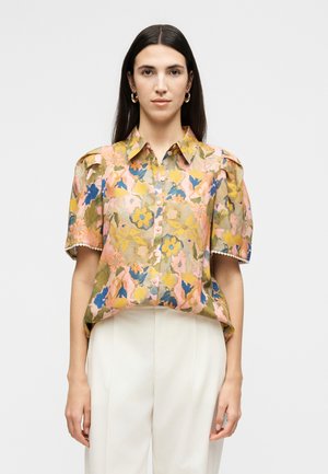 Woman with long dark hair wears floral short-sleeve button-up shirt with puffed shoulders and white high-waist trousers, standing against white background.