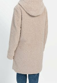 Manteau en polaire beige avec une capuche, présentant un extérieur texturé et doux. Le manteau a des manches longues et arrive au milieu de la cuisse, associé à un jean bleu.
