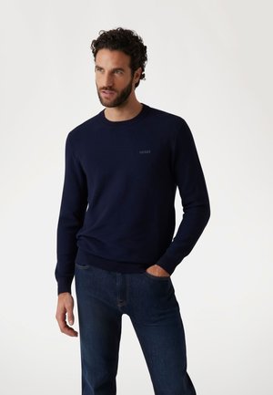 Marineblauer Strickpullover mit Rundhalsausschnitt und gerippten Bündchen. Dezentes Logo auf der Brust, getragen mit dunkelblauen Jeans, die Hände in den Taschen.
