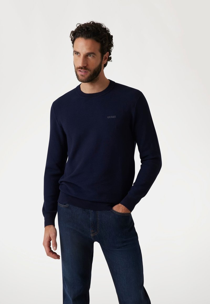 Maglione lavorato a maglia blu navy con collo a giro e polsini a costine. Logo discreto sul petto, indossato con jeans blu scuro, mani nelle tasche.