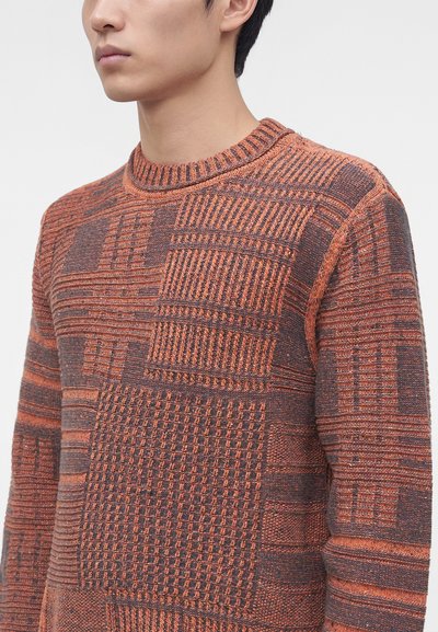 Homme portant un pull en maille texturée avec des motifs géométriques orange et marron, visible du cou jusqu'au milieu du torse, sur un fond uni.
