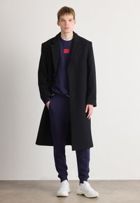 Czarny płaszcz na granatowym sweatshirt'cie z czerwonym logo, w połączeniu z granatowymi spodniami i białymi trampkami. Gładka tekstura, zorganizowany krój.