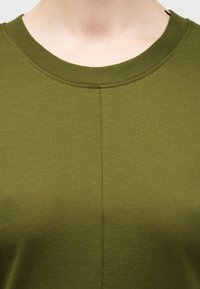 Κοντινό πλάνο ενός ατόμου που φοράει μπλούζα σε στυλ crew neck ελιάς με ένα κάθετο ράψιμο στο κέντρο του μπροστινού μέρους.