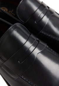 Mocasines de cuero negro con una textura suave, que presentan un detalle de tira frontal y sutiles acentos de costura. La vista en primer plano resalta la artesanía.