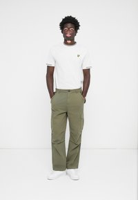T-shirt blanc avec des bordures grises et un petit logo brodé, associé à un pantalon cargo vert olive et des baskets blanches.